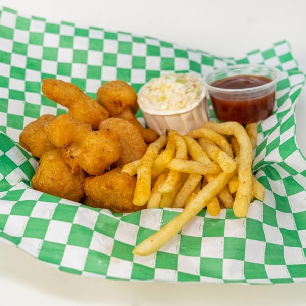 A basket of golden-brown chicken nuggets, french fries, coleslaw, and barbecue sauce on green checkered paper.