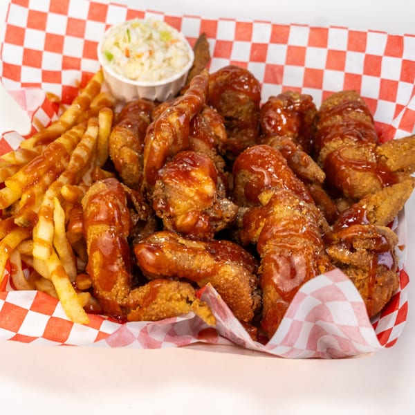 Basket of barbecue-sauced fried chicken wings and tenders with fries and coleslaw on red-and-white checkered paper.