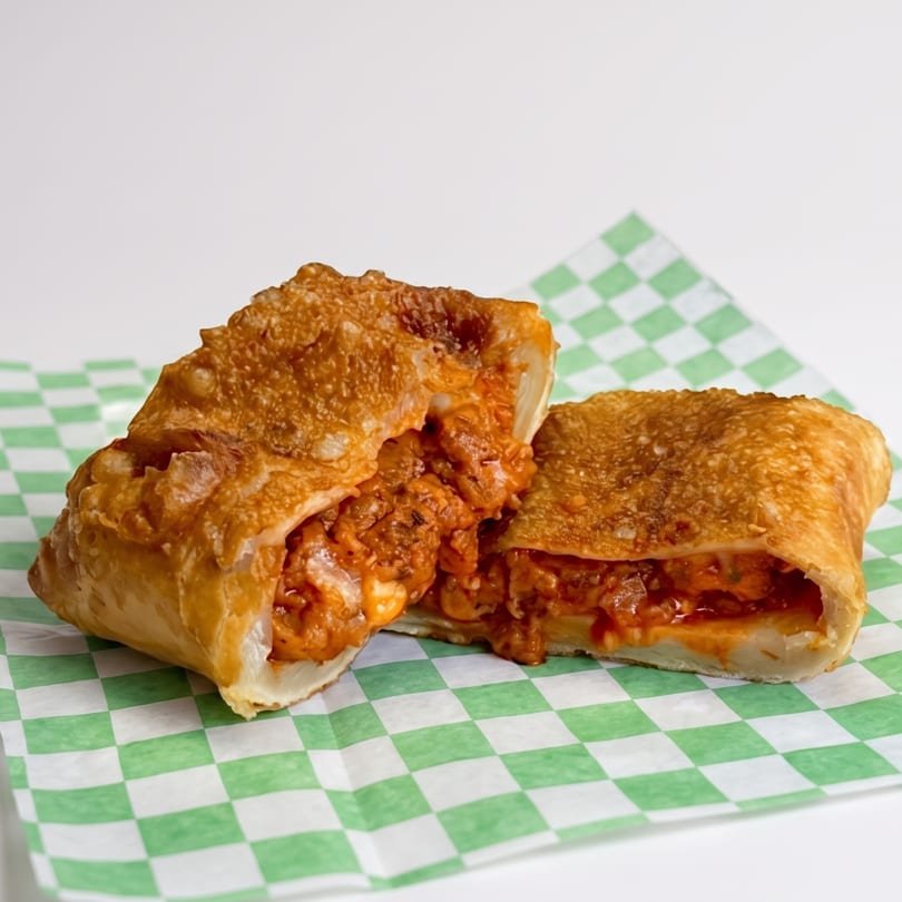 A golden-brown, crispy egg roll, cut in half, revealing a savory, cheesy, orange filling on checkered paper.