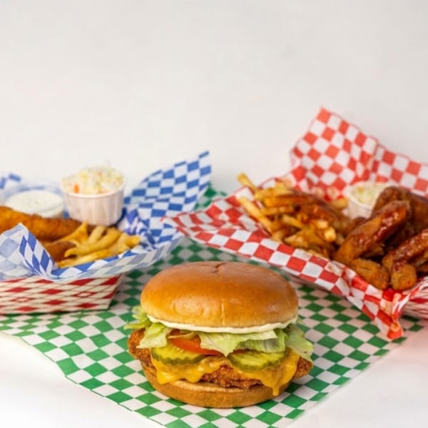 Cheeseburger with lettuce, tomato, pickles, and crispy chicken served alongside fries, wings, dipping sauces, and colorful checkered liners.
