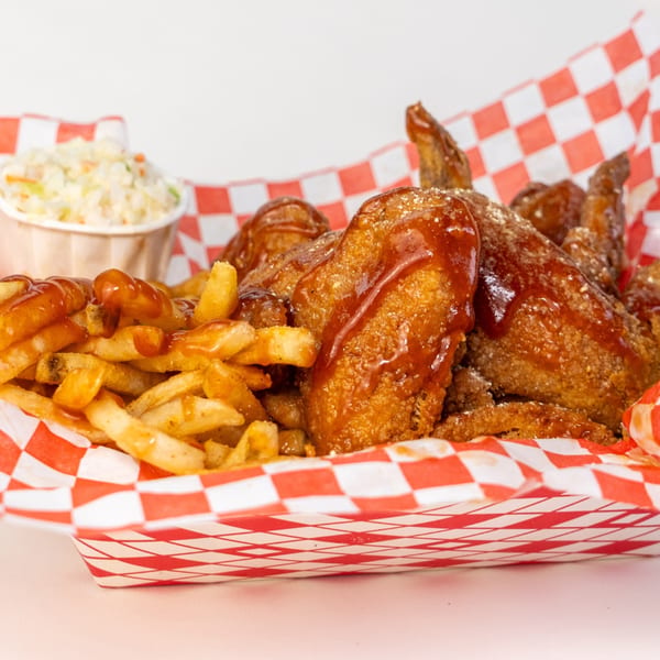 Barbecue chicken wings and french fries with a side of coleslaw in a red and white checkered basket.